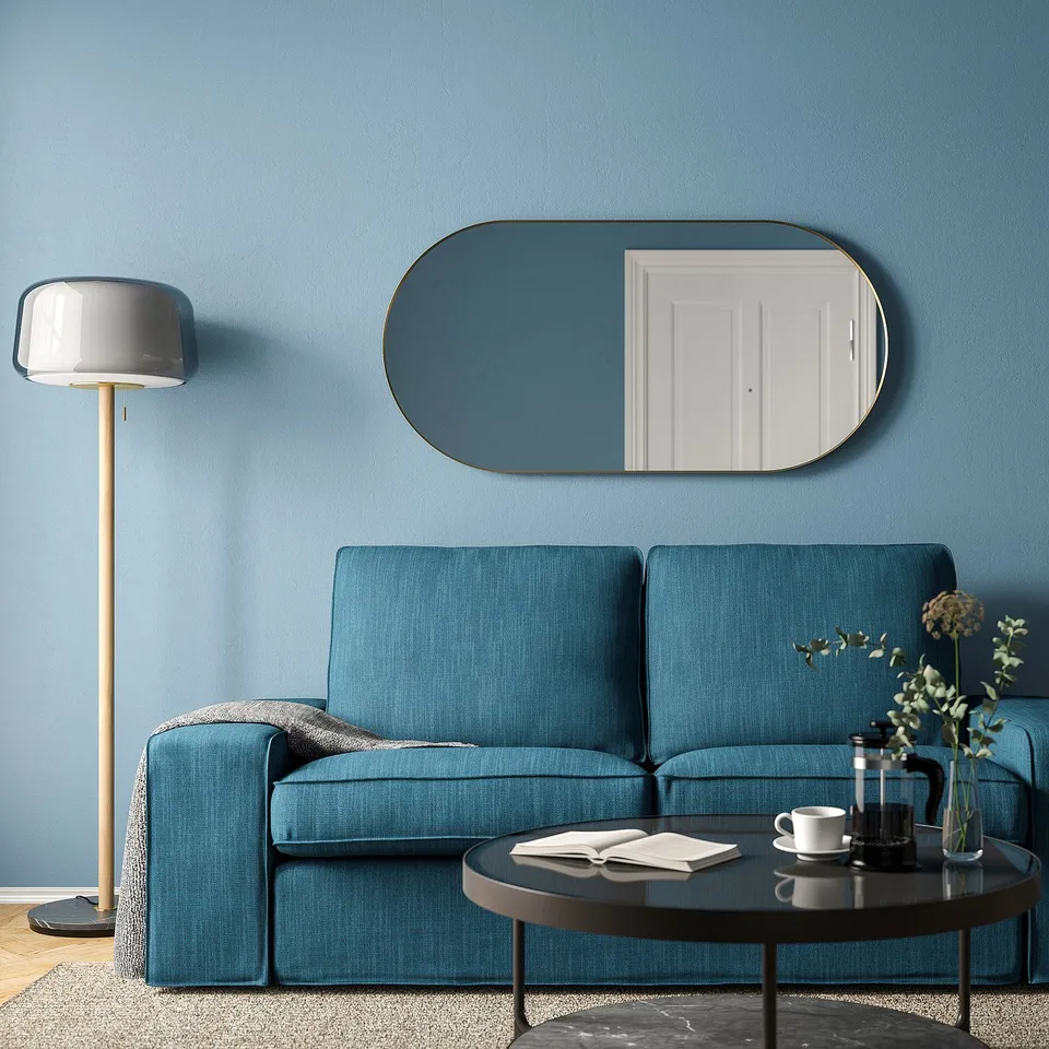 Modern living room with large blue couch, oval gold-framed mirror, stylish floor lamp, and coffee table with cups and plants. Walls are painted blue.