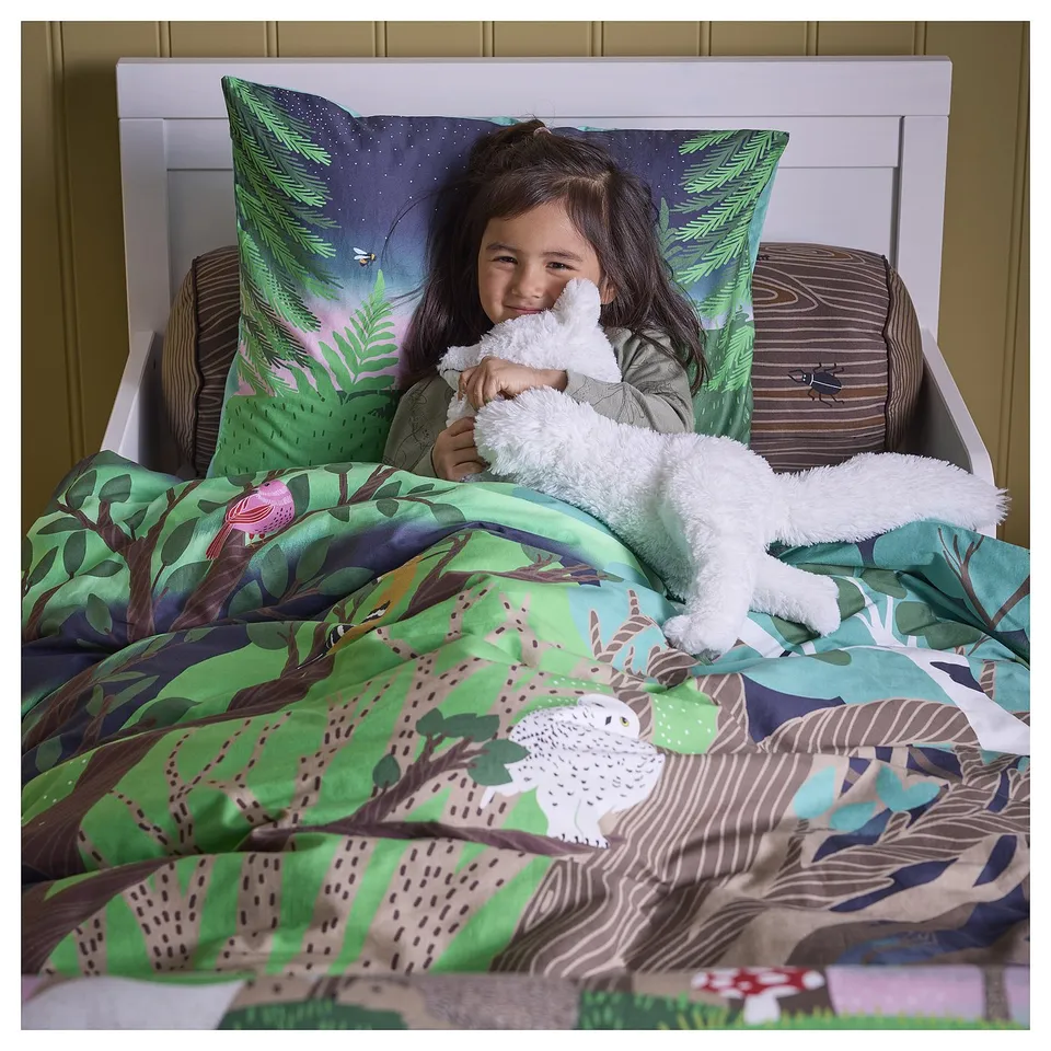 A smiling child hugs a white toy in bed with a green and blue forest-themed duvet and pillows.