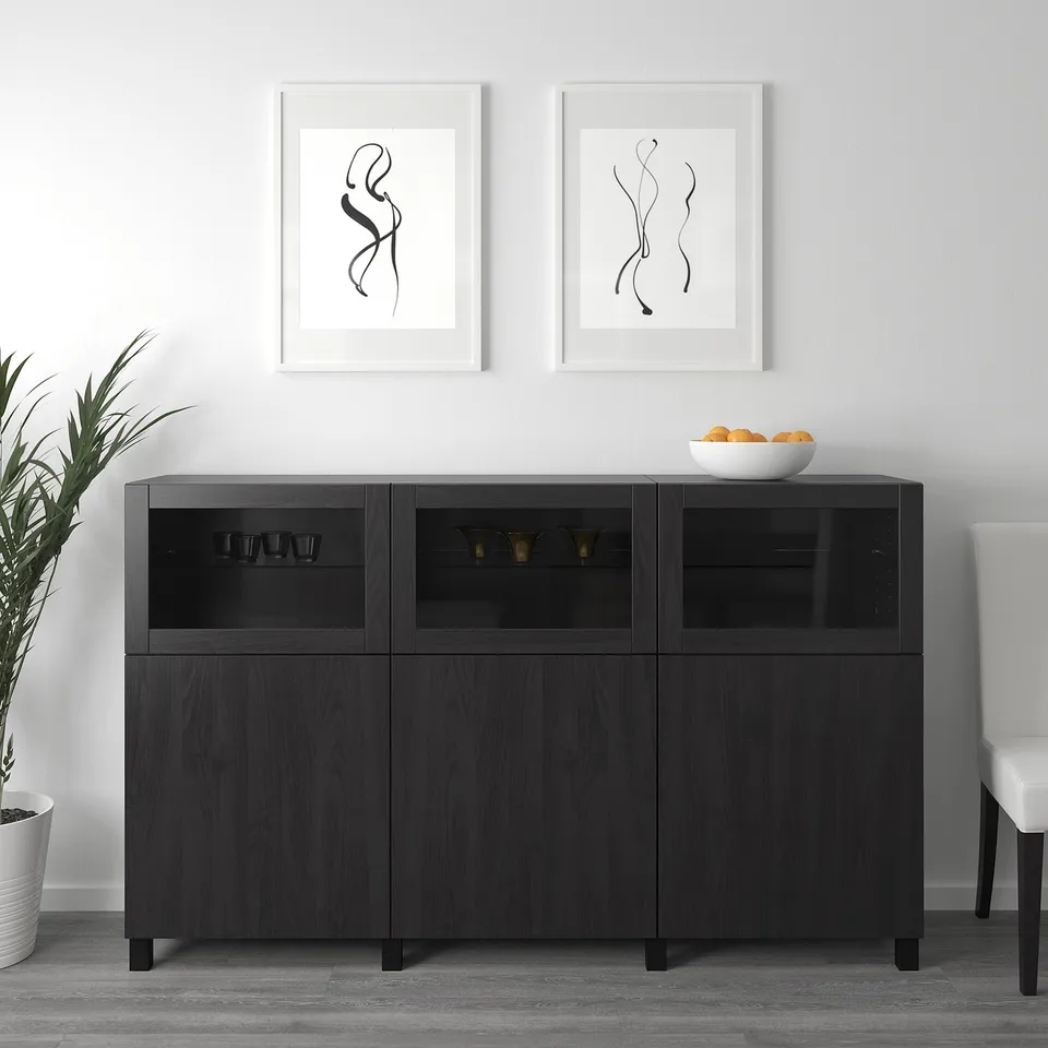Modern black sideboard with glass doors displaying cups and vases. Two framed abstract artworks above. White bowl of oranges on top.