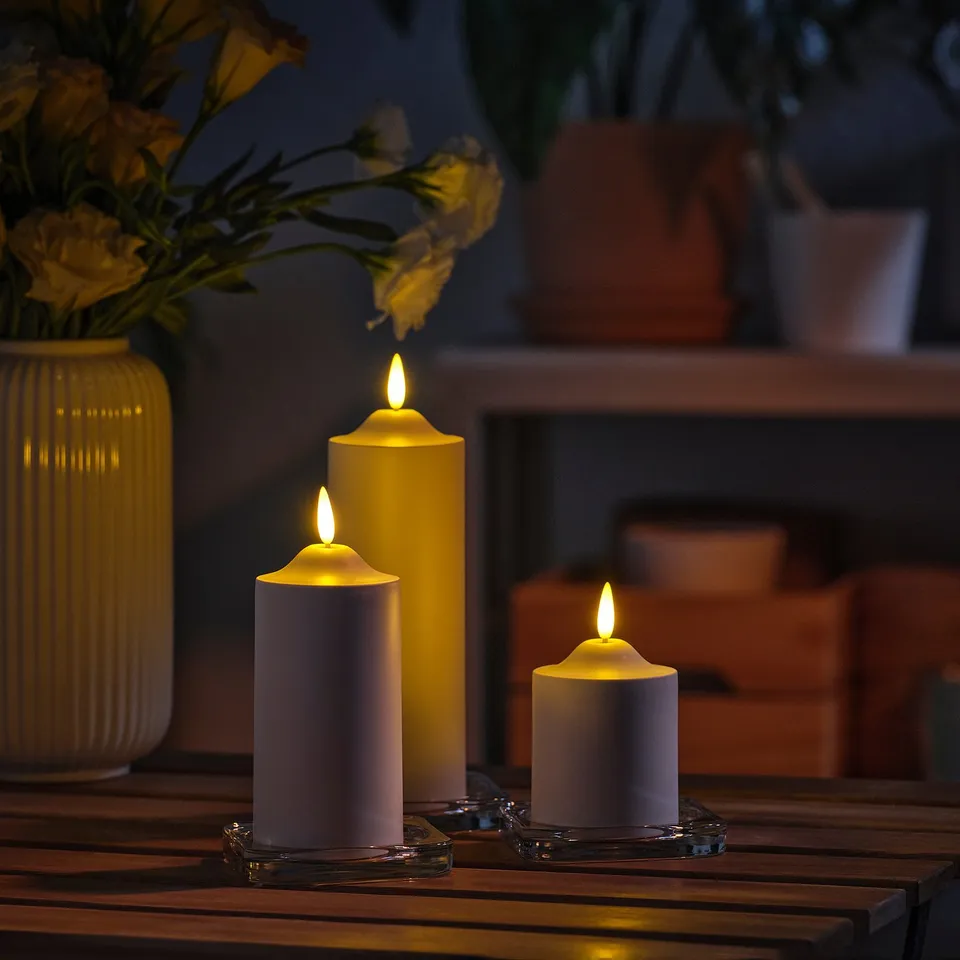 Three flickering led candles in gold and purple, safely in holders, surrounded by flowers in a vase on dark wood.