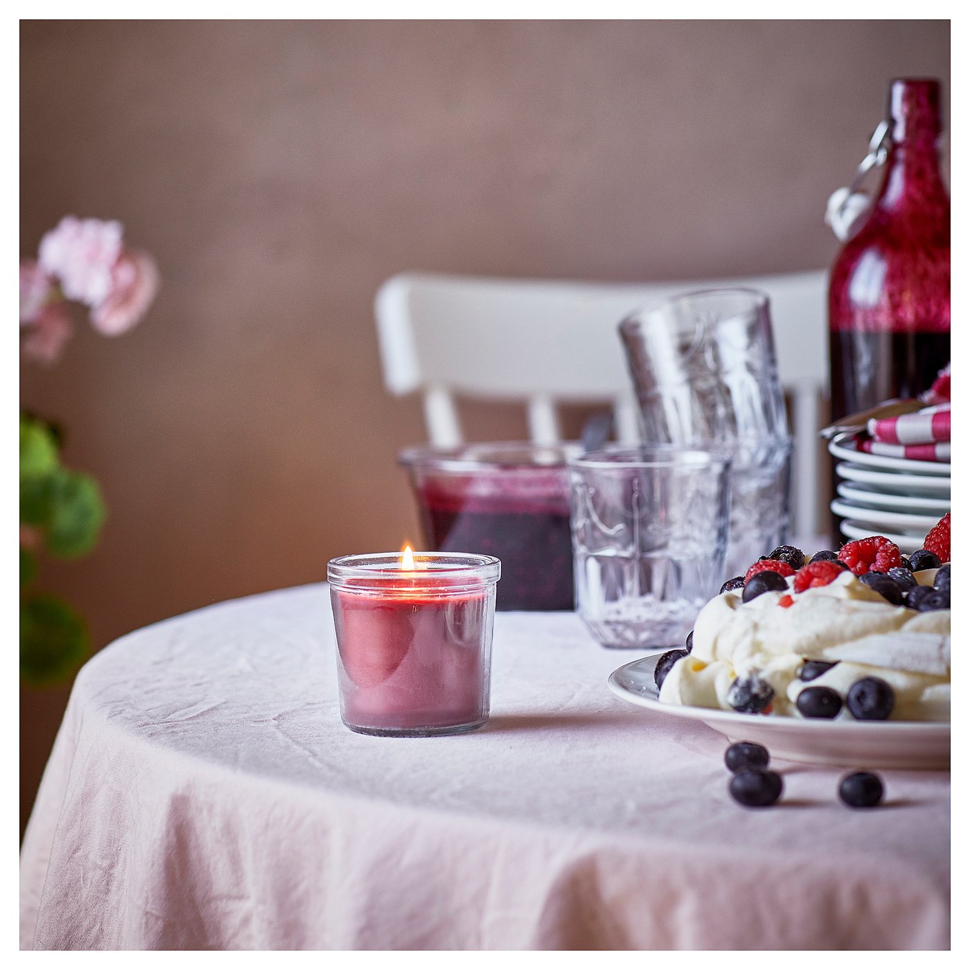 Image a lit red berry-scented candle on a white tablecloth. Accompanied by glasses, a dessert with berries, and vases.