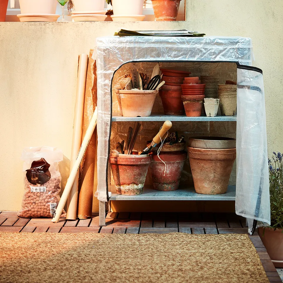 Small indoor/outdoor greenhouse with HYLLIS shelving, terracotta pots, and various plants under a light blue cover.