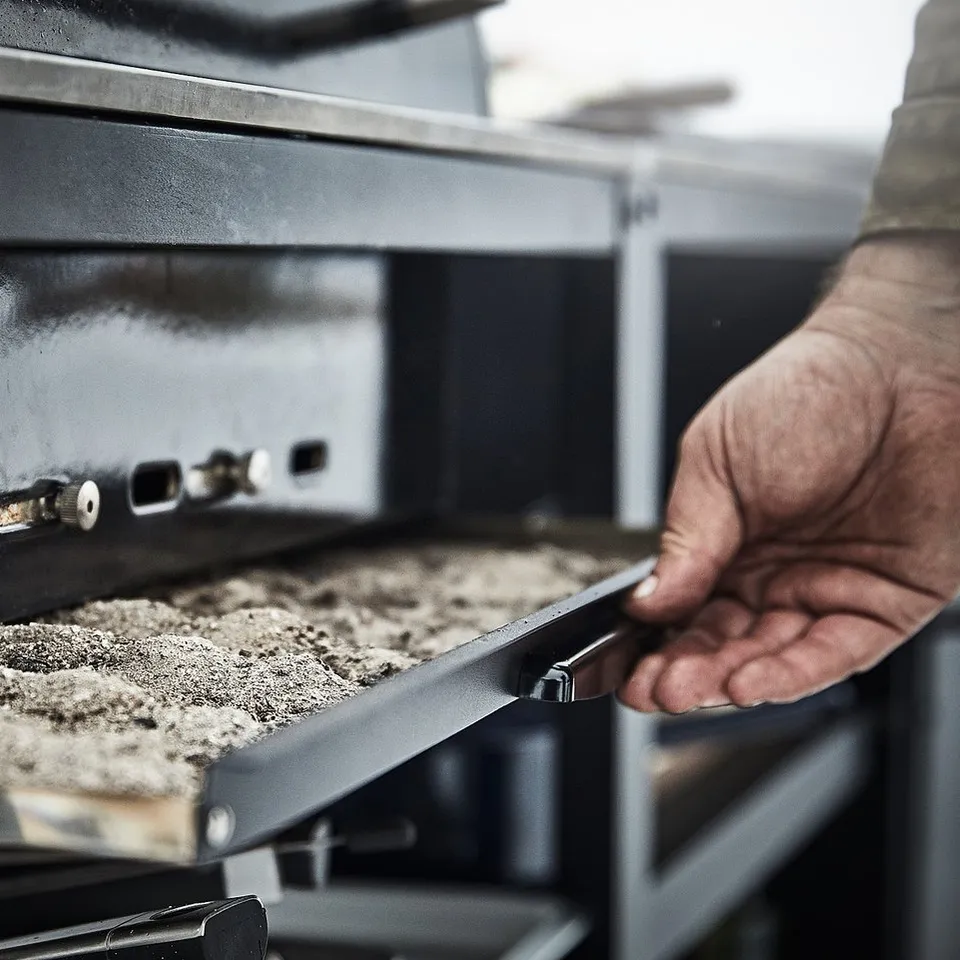 Person opening black GRILLSKÄR charcoal grill, revealing inside with ash tray.