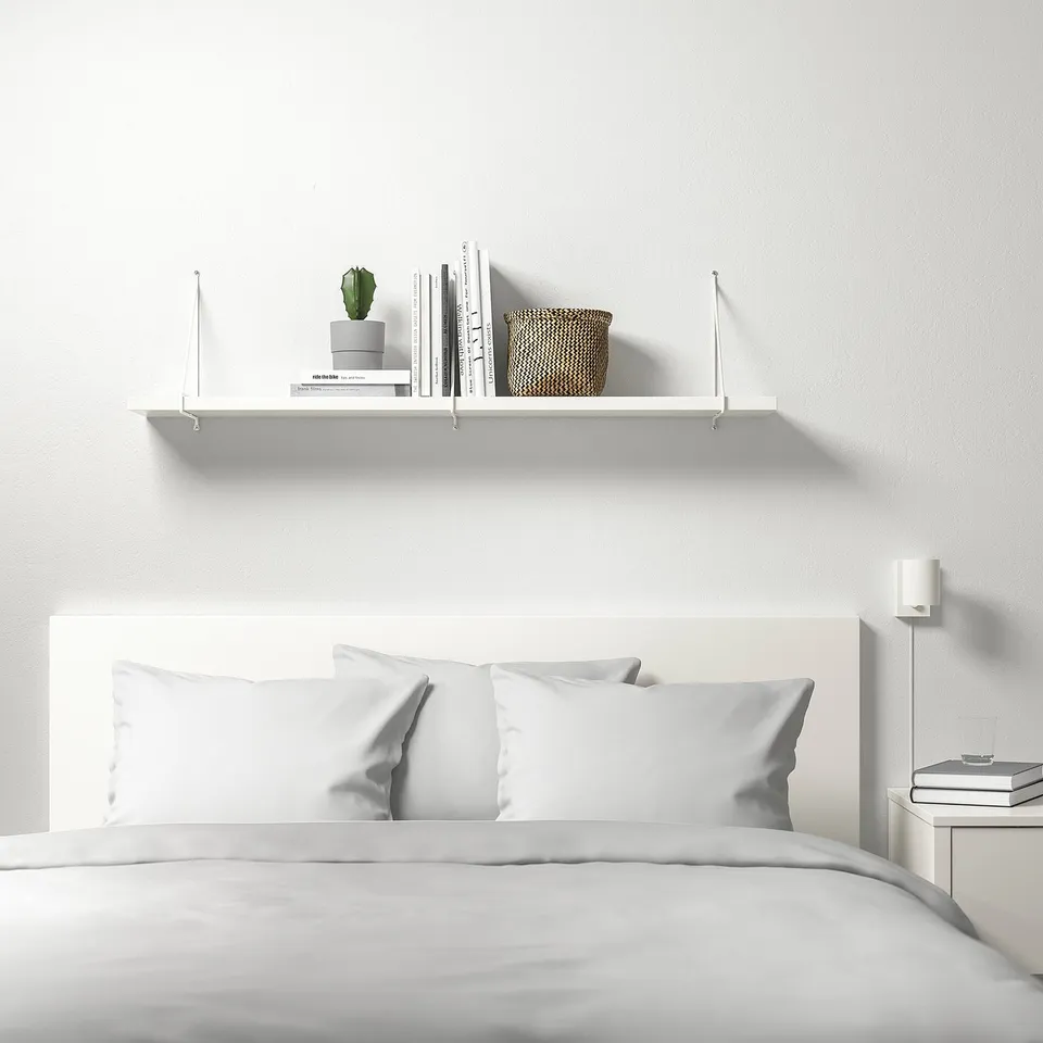 Minimalist bedroom with white BERGSHULT shelf above bed, holding plant, books, and basket. Bed has white pillows and headboard.