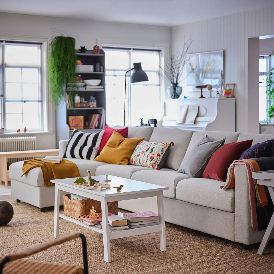 Bright living room with VIMLE sofa, colourful pillows, white coffee table with magazines, rug, and large windows.