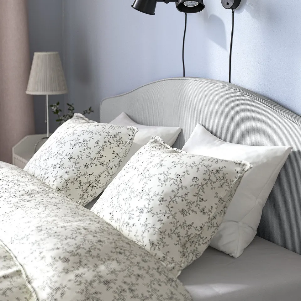 Bedroom scene: a cosy upholstered bed with a curved headboard, white patterned pillows, and a bedside lamp.
