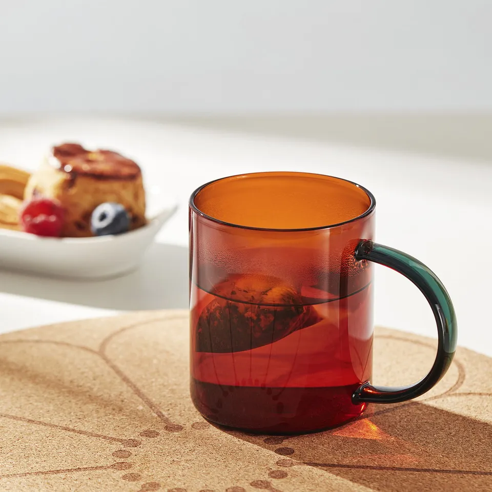 A red glass mug with a green handle sits on a cork mat, showcasing its comfortable grip and stylish design.