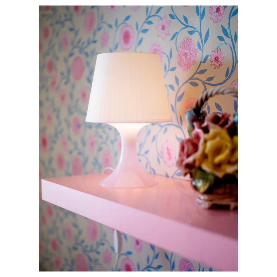 A white lamp on a pink shelf, casting a soft glow, with floral wallpaper background.