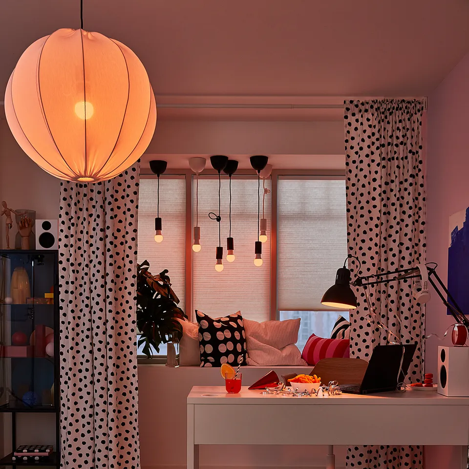 Modern office space with a MICKE desk, black and white polka dot curtains, striped rug, hanging lights, laptop, and speakers, in soft pink tones.