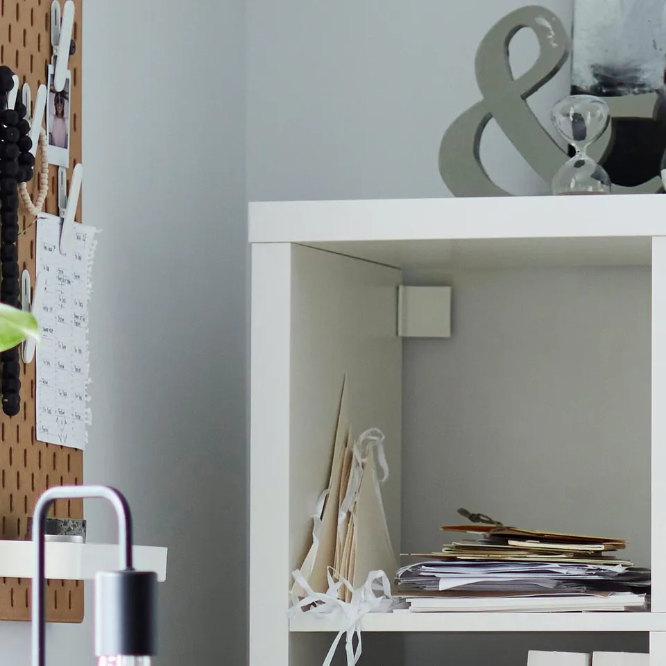 White KALLAX shelving unit with clear boxes and scattered papers. A pegboard with notes and a lamp nearby.