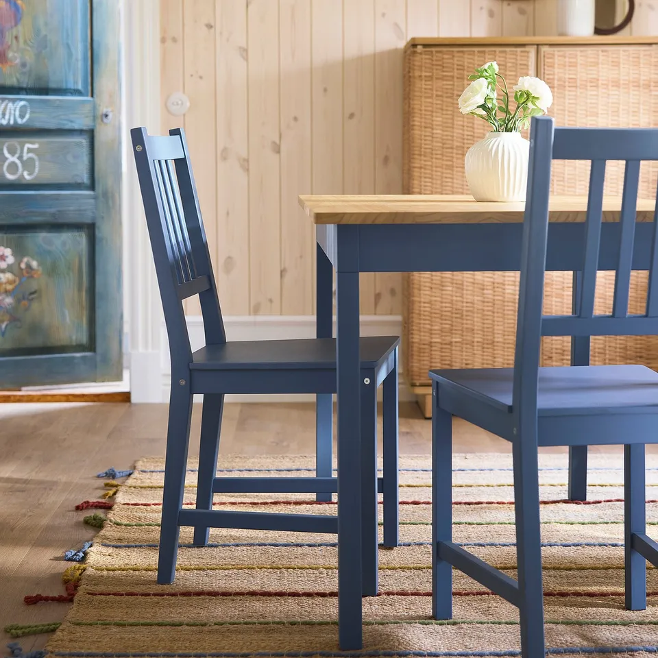 Two dark blue wooden chairs near table with white vase, against wooden wall.