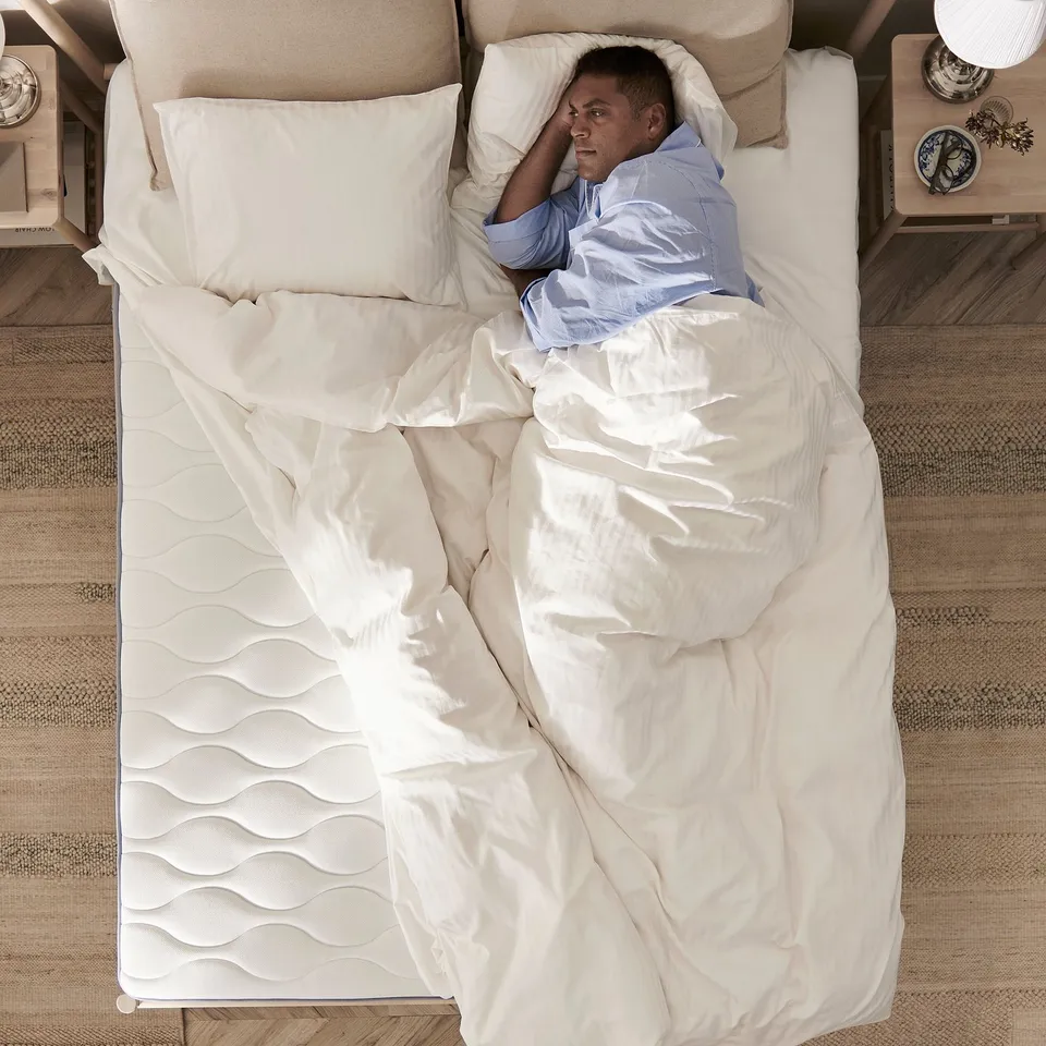 Person sleeps on firm, white VÅGSTRANDA mattress in cosy bedroom.