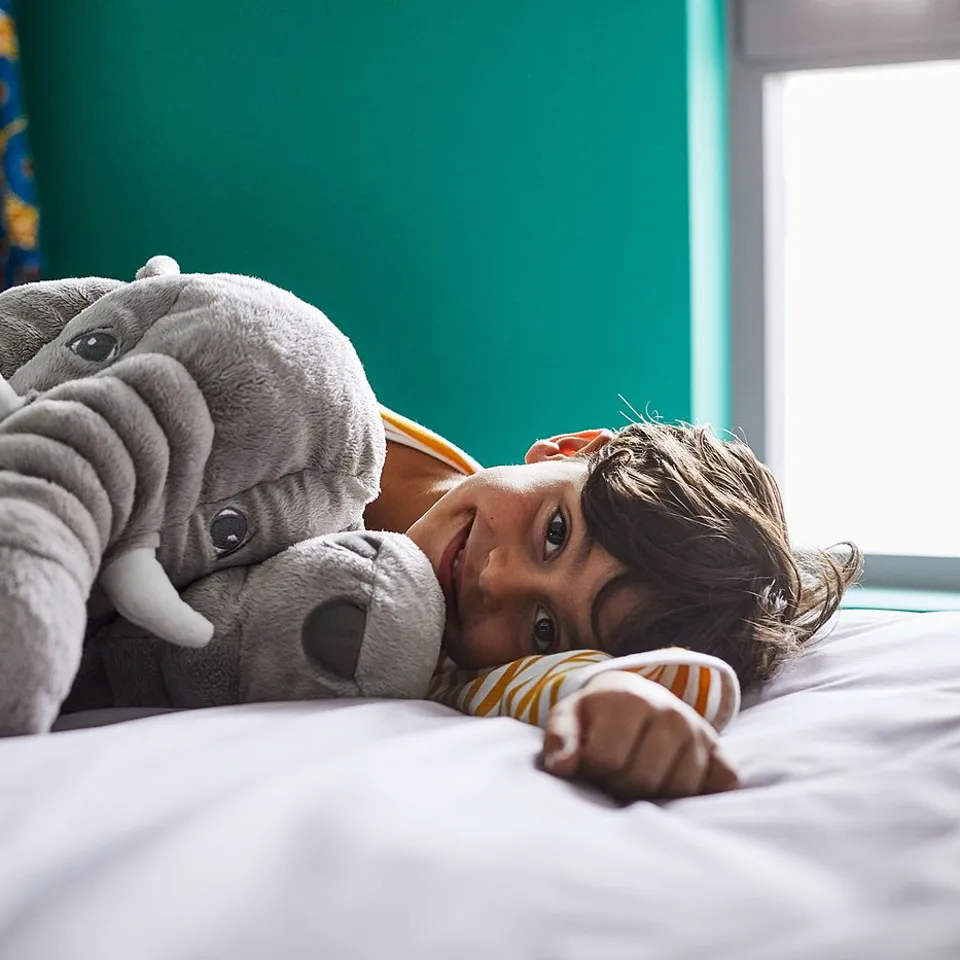 A child hugs a large grey elephant cuddly toy on a bed. The toy is soft with white tusks and floppy ears.