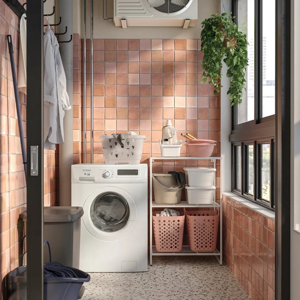 Small laundry room with white JOSTEIN storage shelves, pink bins, and hanging plants.
