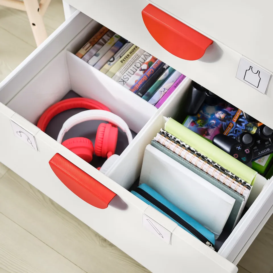 White SMÅSTAD kids storage drawers with red handles, storing headphones, books, and notebooks in a neat arrangement.
