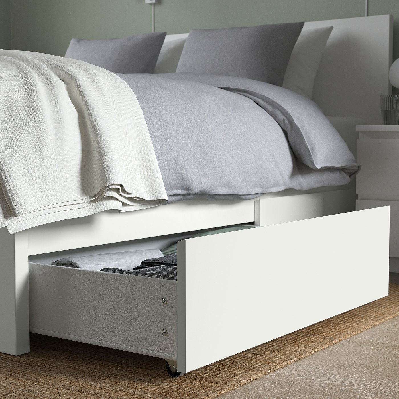 A white MALM bed with a rolled-out storage drawer filled with blankets. The bed has a clean design suitable for any bedroom.
