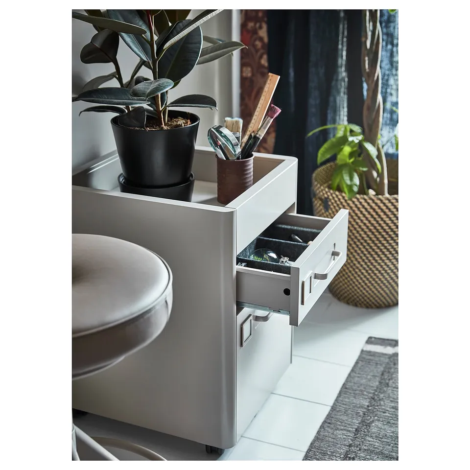 An IDÅSEN silver drawer unit with a potted plant and office supplies on top. The drawers are partially open, revealing an organised interior.
