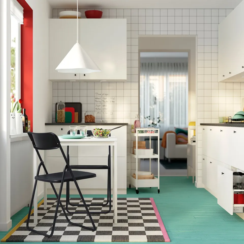 Kitchen with white cabinets, turquoise floor, black-and-white rug. Red accent wall, small table, black chair. Bright lighting.