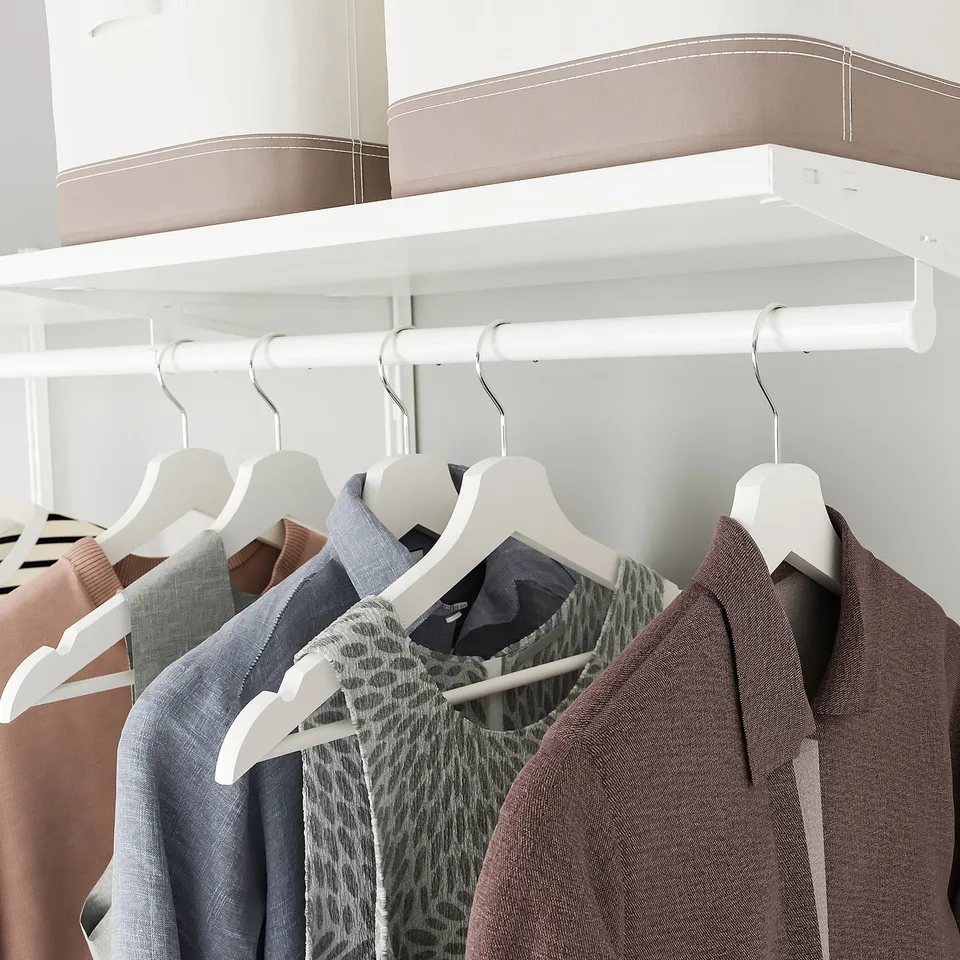 A closet with white SKYTTA rail holding clothes on hangers and a shelf with storage boxes above.