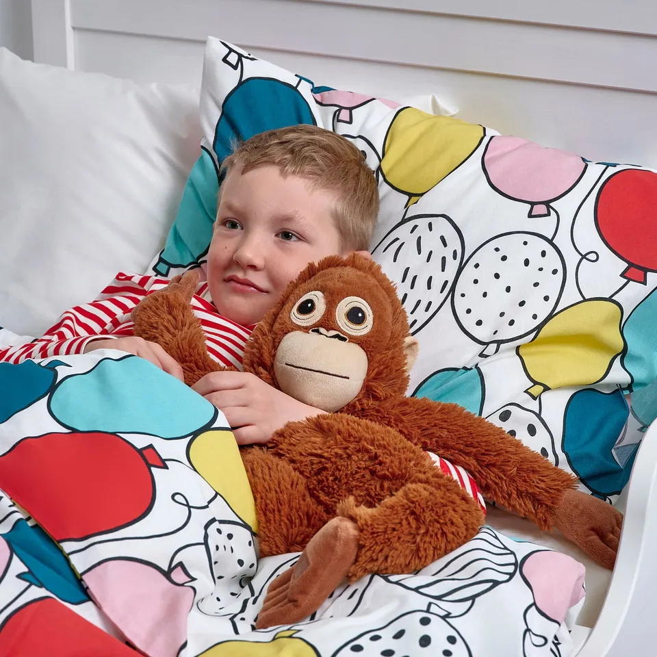 Child hugging soft orange monkey toy in bed, with colourful pillows.