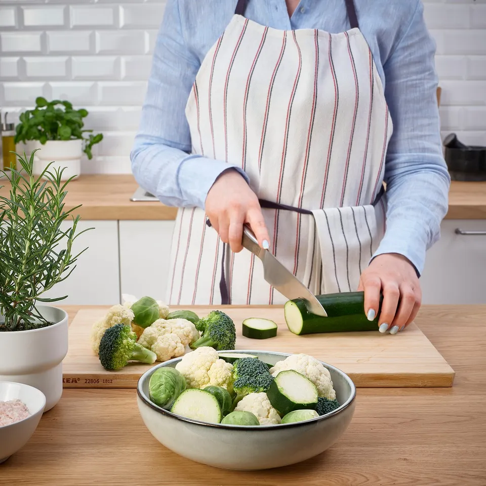 Person slicing courgette into bowl of veggies on wooden cutting board with oven dish nearby.