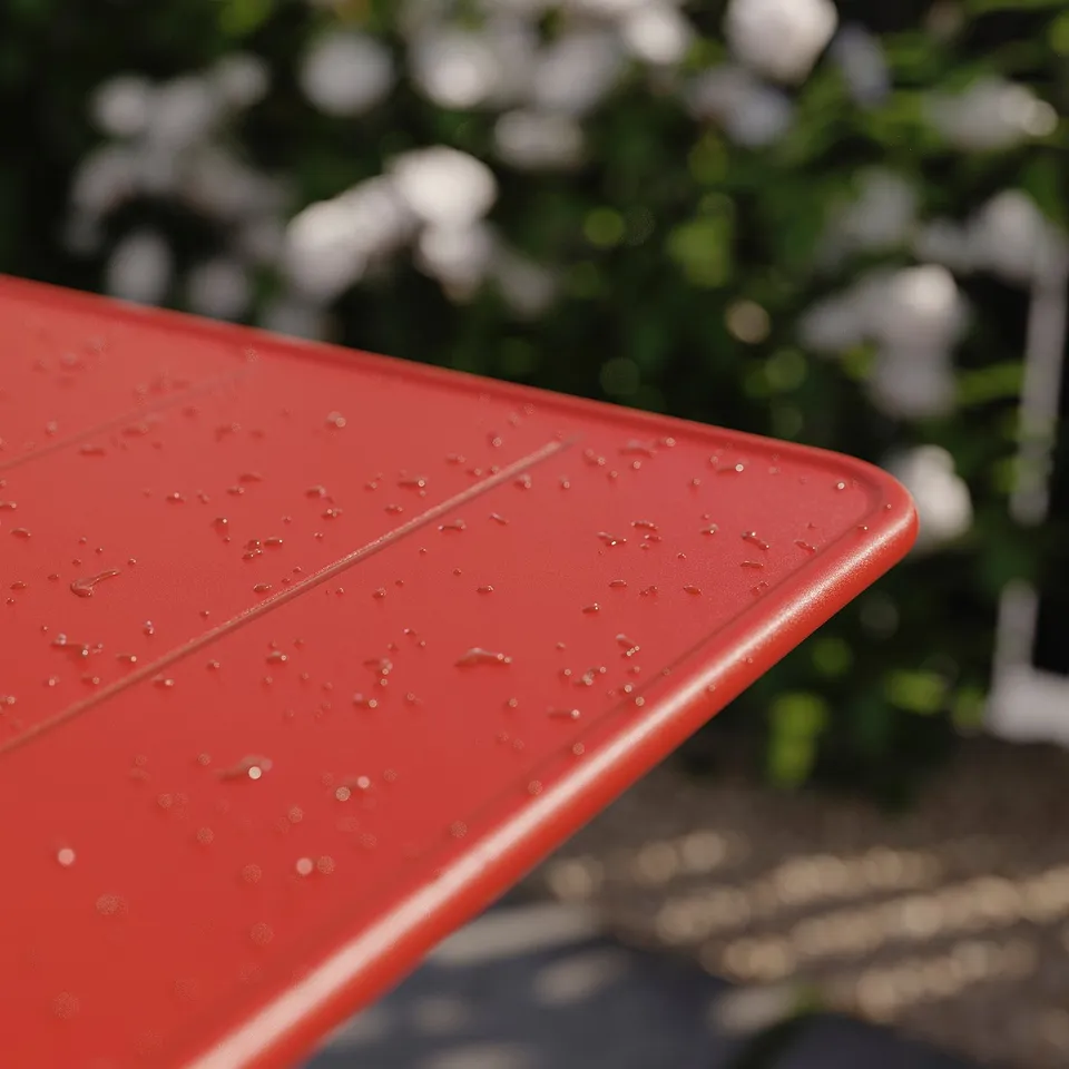 Red plastic outdoor table top with water droplets, rounded edges, in front of green plants.