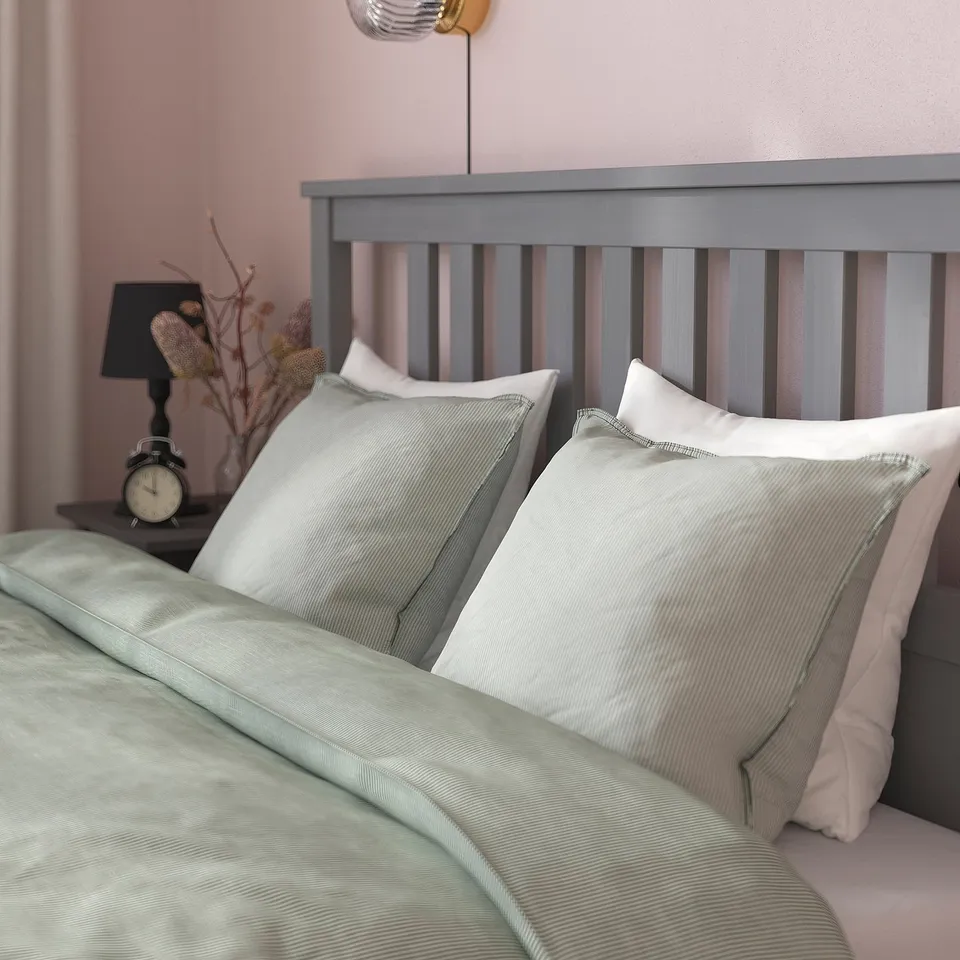A cosy bed with a grey wooden headboard, white pillows, and a green blanket. An alarm clock and dried flowers sit on the bedside table beside it.