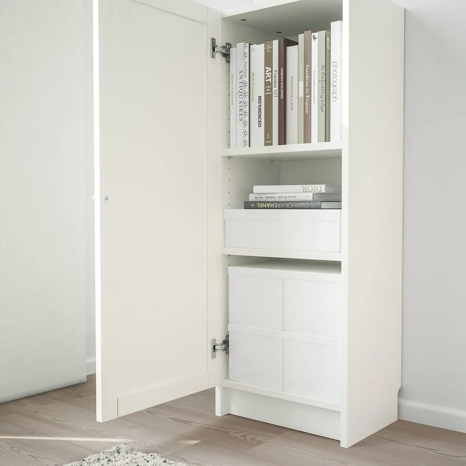 White bookshelf with closed door, housing books and boxes in organised manner.