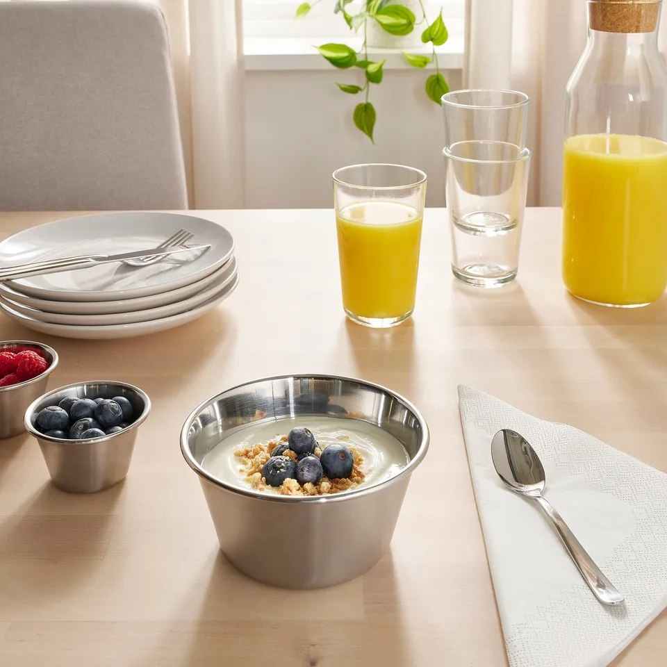 Table set with stainless steel bowl of yoghurt, berries, and granola. Glasses of orange juice and water, plates, cutlery, and a plant.