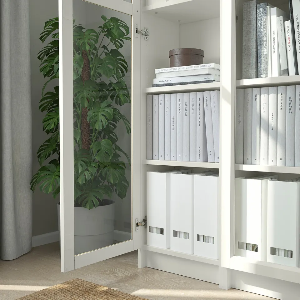 White BILLY bookcase with glass doors, showcasing books and a potted plant, reflecting a cosy, organised space with natural light.