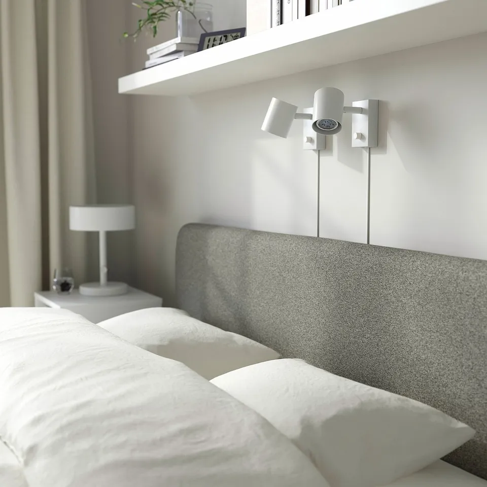 Bedroom scene with SLATTUM bed, white pillows, grey headboard, wall-mounted light, bedside table with lamp.