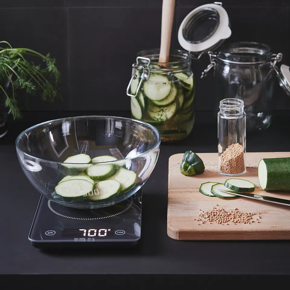 Kitchen setup: glass bowl with sliced cucumbers on scale, jars and cutting board with seeds and knife, dark backdrop.