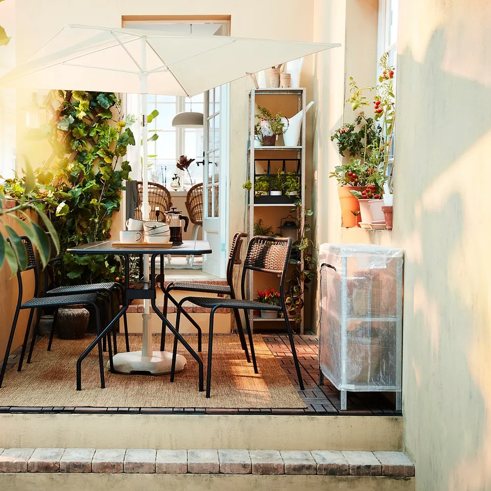 Patio with black table, chairs, green plants, and white umbrella. Right: plant storage unit.