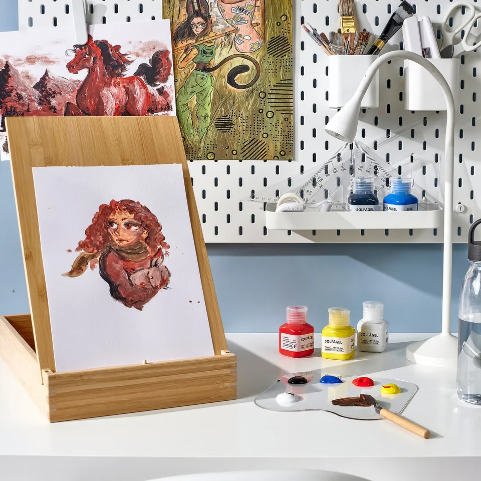 Art studio with SOLFÅGEL easel, acrylic portrait, and supplies on desk.