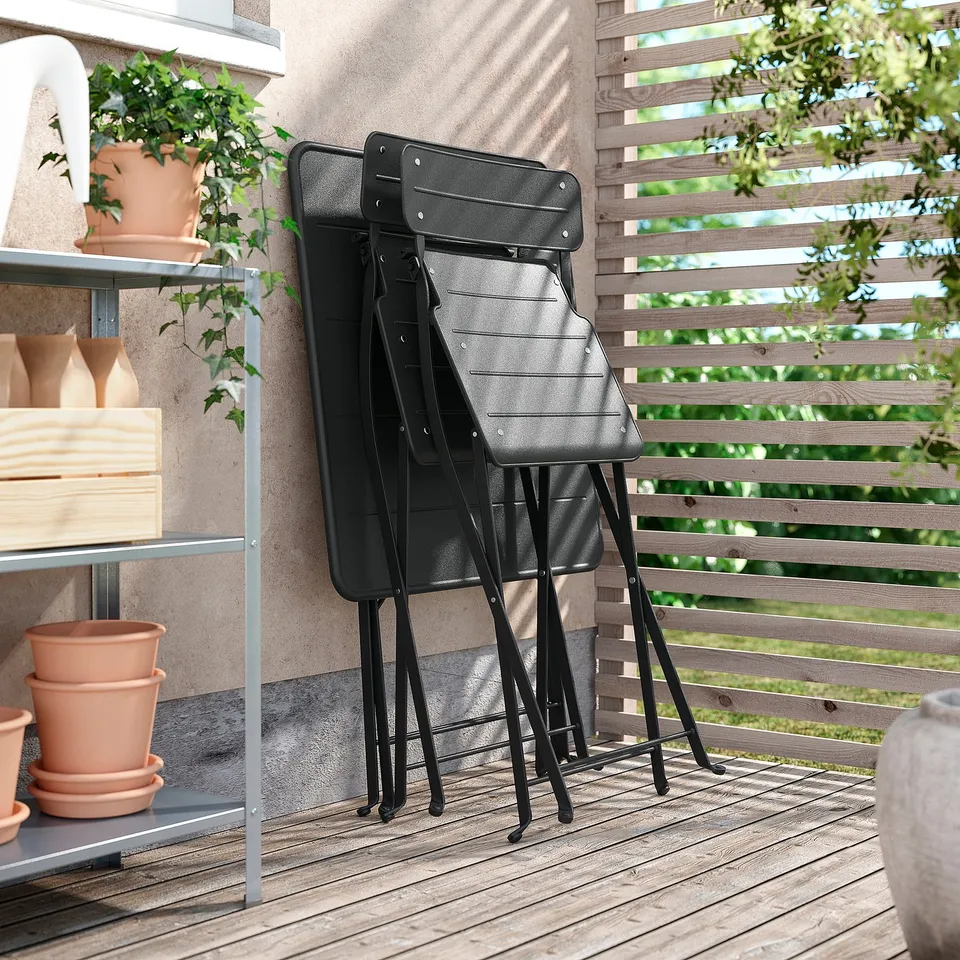 Black folding chairs stacked outdoors, shown with wooden fence and potted plants.