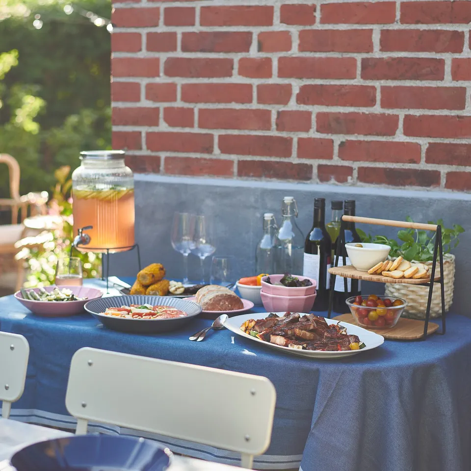 Outdoor table with blue tablecloth, various foods, drinks, and a VARDAGEN jar stand with lemonade. Red brick wall background.