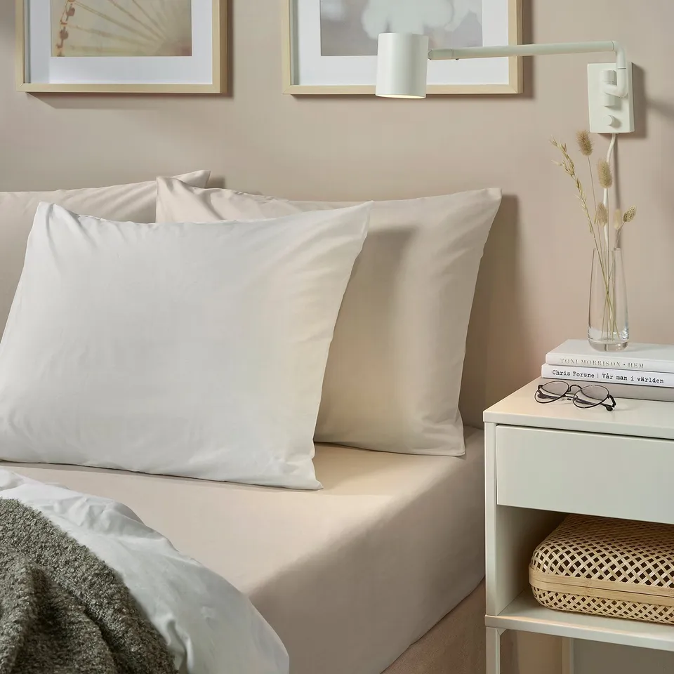 Bed with large white DVALA pillows, light beige bedding, small bedside table holding books, glasses, and a clear vase with dried flowers.