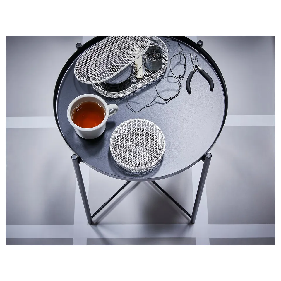 A GLADOM side table in grey. It has a round glass top and a metal mesh tray witharlike tools, a mug, and a cup of tea.