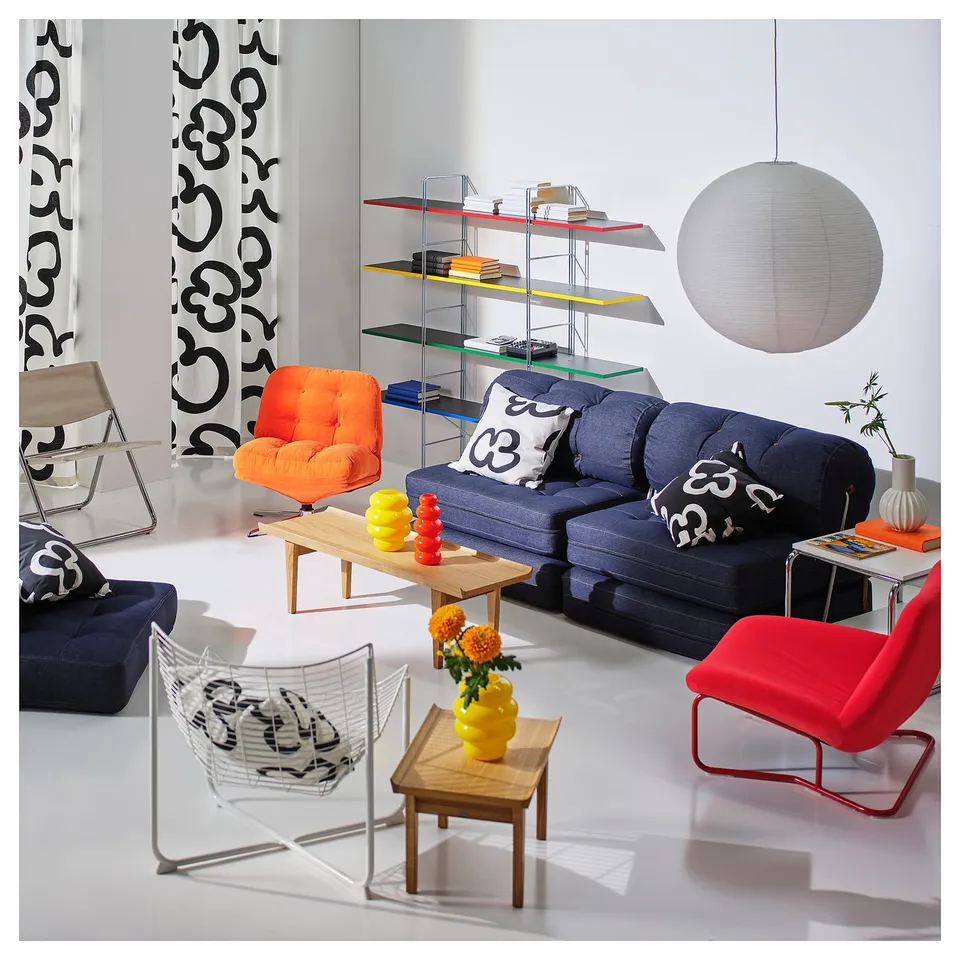 Bright living room: blue couch, orange/red chairs, large white light, glass shelves, yellow vase, patterned curtains.