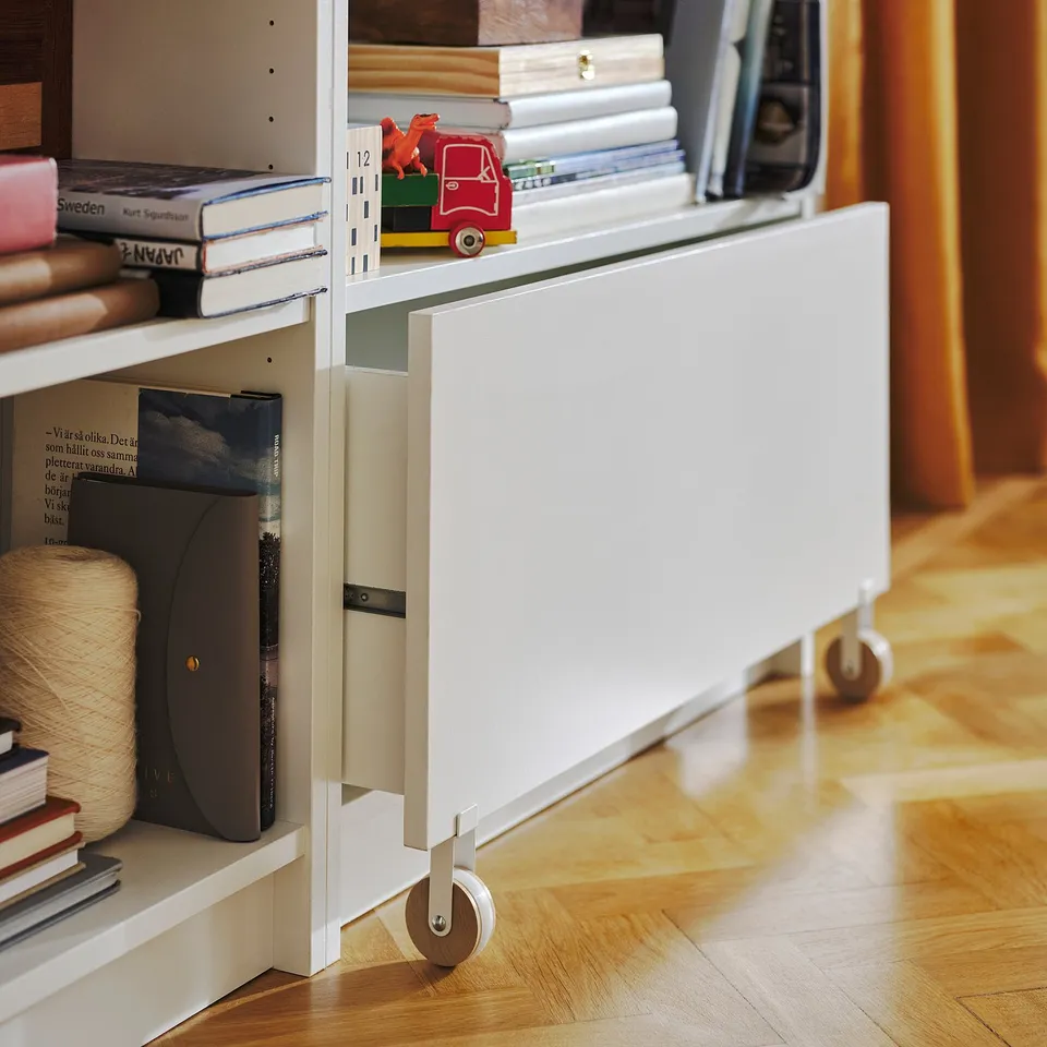 White BILLY bookshelf with drawers, wooden wheels, holding books, toy car, wooden floor.