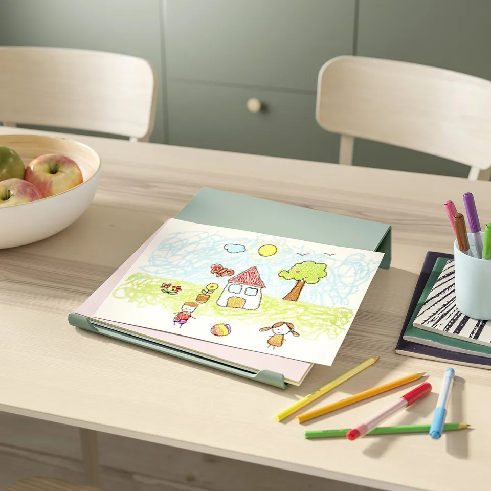 Childs drawing on light green writing board, pencils, bowls of apples on light wooden table with two white chairs.