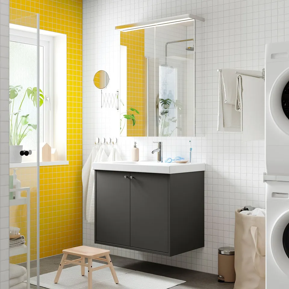 Grey vanity, white counter, yellow tiles, mirror, towels, washing machine, stool.