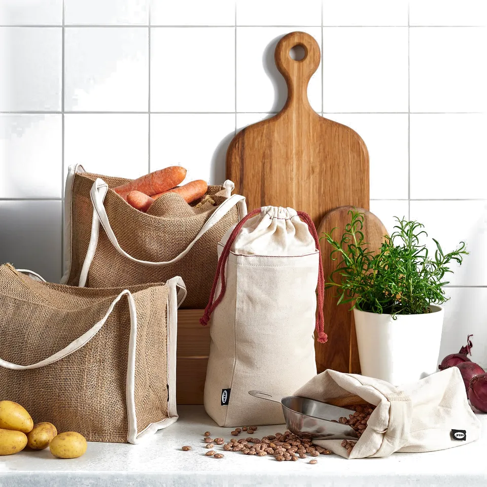 Beige jute bags with white handles store veggies, cutting boards, and herbs on kitchen counter.