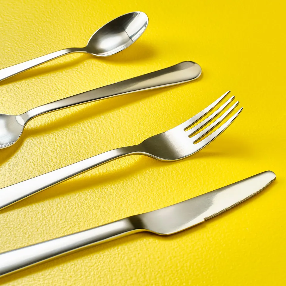 Polished stainless-steel knife, fork, spoon on yellow background, reflecting light.