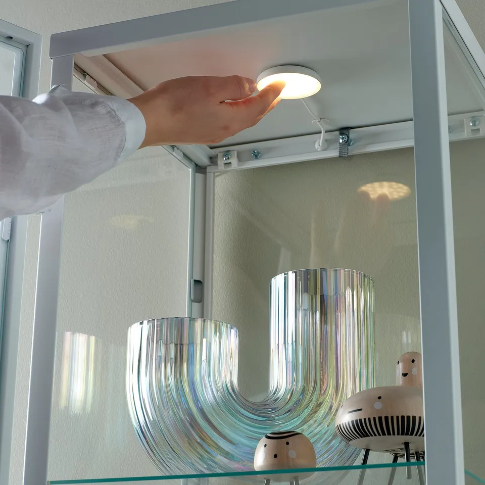 A hand turns on a small cabinet light, illuminating decorative items inside a glass-fronted cabinet.