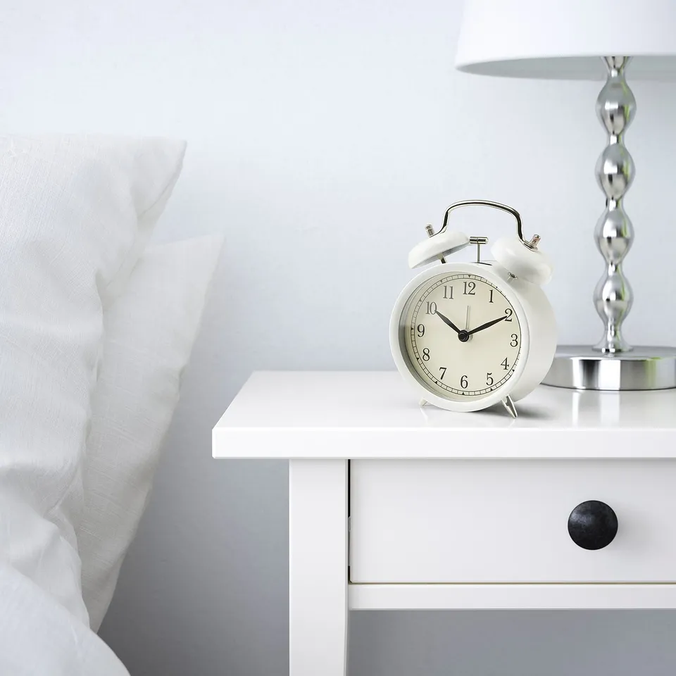 White bedside clock with silent quartz movement sits on a white bedside table. Next to it, a lamp and white pillow. Display shows 10:10.