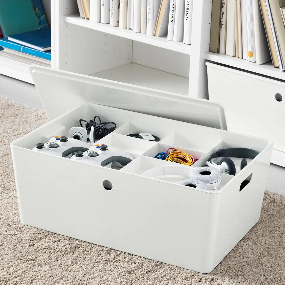White organiser box with multiple compartments holding game controllers and cables, placed on rug near bookshelf.