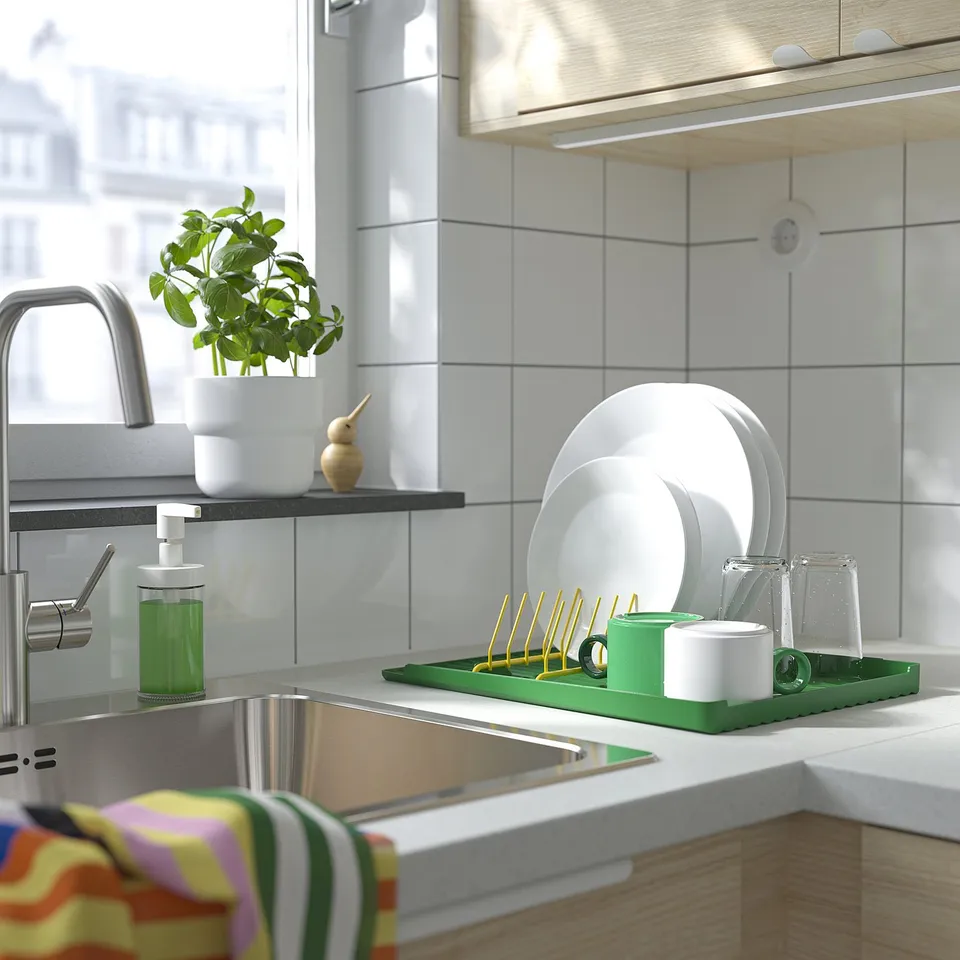 Modern kitchen sink with stainless steel tap, green dish drainer, white dishes, plant, and colourful towels.