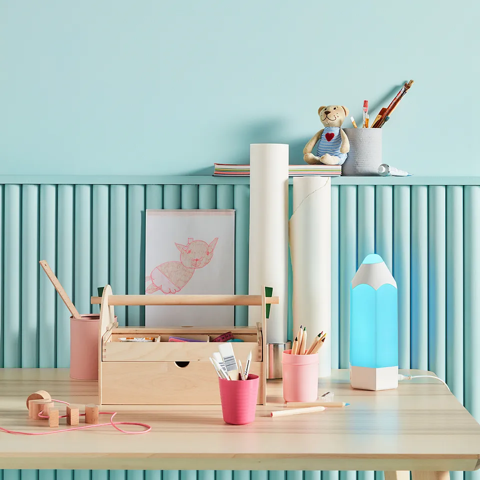 Kids desk with blue light, art supplies, teddy bear. Pink cups, wooden toys. Light blue walls, vertical lines.