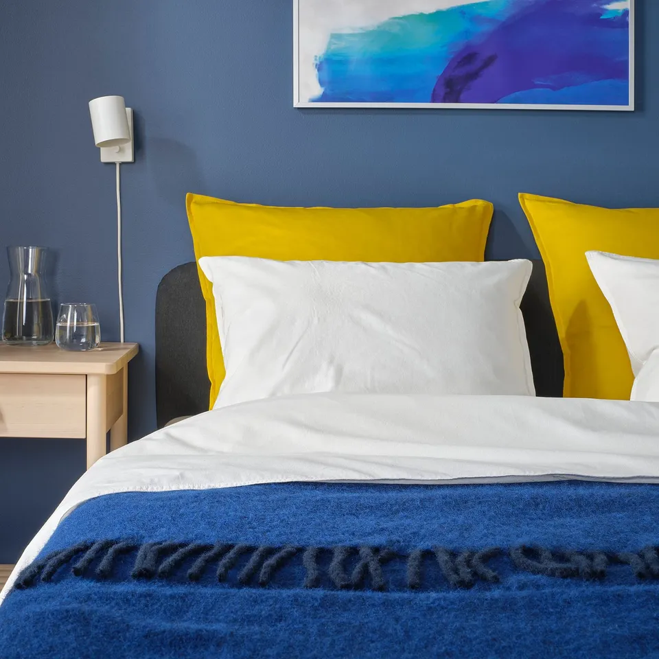 Bedroom with yellow GURLI cushion on a bed, blue walls, and abstract wall art.